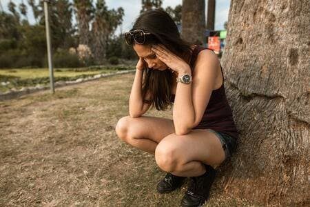 Foto de uma mulher em um parque com shorts e regata pretas. Ela está agachada com as mãos na cabeça, enconstada em uma árvore, como se estivesse tentando se acalmar de um ataque de ansiedade. A foto ilustra o artigo "Psicanalistas que tratam a Ansiedade Desvendam Tabus e Mudam Vidas" escrito por Homero Mônaco para o site Online Psicanalista.