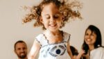 Foto de uma menina de cabelo castanho claro enrolado em primeiro plano, pulando na cama. Em segundo plano aparece o pai, ao lado da mãe com um bebê no colo. Ambos estão sorrindo. A foto ilustra o artigo "Revelando o poder das experiências da infância" escrito pelo psicanalista Homero Mônaco para o site Online Psicanalista.