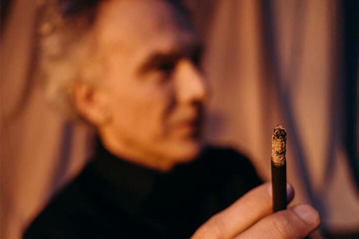 Foto de um homem de perfil, sentado e em primeiro plano está a mão dele com uma cigarrilha na mão. A foto ilustra o artigo 7 maneiras de como a psicanálise vai te ajudar no tratamento da dependência escrito no blog do psicanalista Homero Mônaco para ao site Online Psicanalista.Foto em tons azuis do rosto de perfil de uma jovem com a boca aberta, olhos fechados e um comprimido em sua língua. A foto ilustra o artigo 7 maneiras de como a psicanálise vai te ajudar no tratamento da dependência escrito no blog do psicanalista Homero Mônaco para ao site Online Psicanalista.
