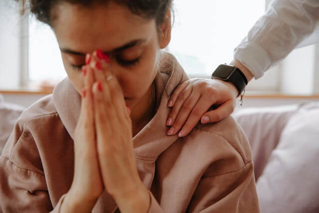 Foto de umuma mulher com as mãos juntas no rosto e cara de preocupada. É possível ver a mão de uma pessoa em seu ombro como se estivesse usando a empatia para dar à outra mulher algum apoio. A foto ilustra o artigo 10 Benefícios da Terapia para a Expansão da sua Consciência Emocional escrito pelo psicanalista Homero Mônaco para o site Online Psicanalista.
