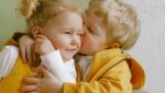 Foto de um menino loiro beijando o rosto de uma menina loira que sorri se divertindo mostrando o seu bem-estar emocional. A foto ilustra o artigo 10 Melhores Práticas para Promover o Bem-Estar Emocional escrito pelo psicanalista Homero Mônaco para o site Online Psicanalista.