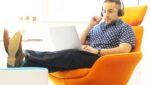 Foto de um homem sentado em uma cadeira laranja, em um escritório, com fone de ouvidos e olhando para o laptop que está em cima das suas pernas. Ele está com as pernas erguidas, com os pés em cima de uma mesa e com roupa informal, representando o equilíbrio entre o trabalho e a vida pessoal. A foto ilustra o artigo 5 Estratégias para Equilibrar Trabalho e Crescimento Pessoal escrito pelo psicanalista Homero Mônaco para o site Online Psicanalista.