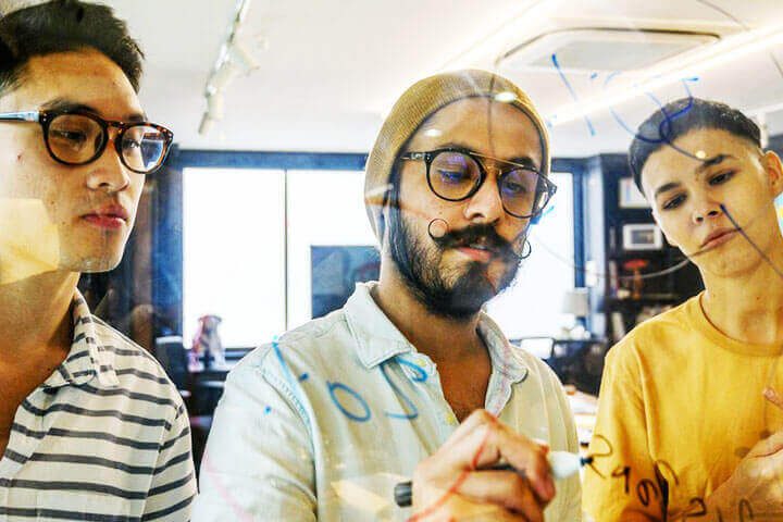 Foto de um homem escrevendo em um vidro sobre metas de crescimento enquanto outros dois observam. A foto ilustra o artigo 5 Estratégias para Equilibrar Trabalho e Crescimento Pessoal escrito pelo psicanalista Homero Mônaco para o site Online Psicanalista.