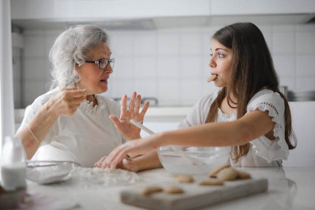 Foto de uma avó sentada na mesa da cozinha com a neta. A avó parece estar a repreendendo porque ela pegou uma das bolachas que fizeram e está com ela na boca. A foto ilustra o artigo Compreenda 80% das suas motivações ocultas por trás das suas ações escrito pelo psicanalista Homero Mônaco para o site Online Psicanalista.