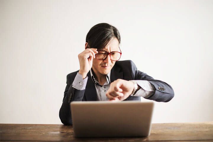 Foto de um homem oriental, de terno, sentado. Na mesa está um laptop, e ele está olhando o seu relógio de pulso como se estivesse fazendo o gerenciamento do seu tempo. A foto ilustra o artigo 5 Estratégias para Equilibrar Trabalho e Crescimento Pessoal escrito pelo psicanalista Homero Mônaco para o site Online Psicanalista.
